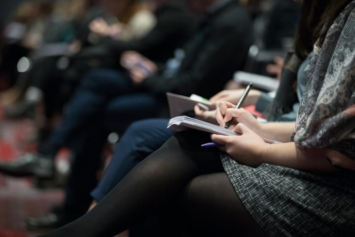 A woman sits among others in a row of chairs, writing in a notepad.
